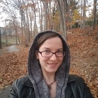 Morgan, dressed in a grey hoodie and black leather jacket stands on a walking trail with trees and a ground covered in leaves behind her.