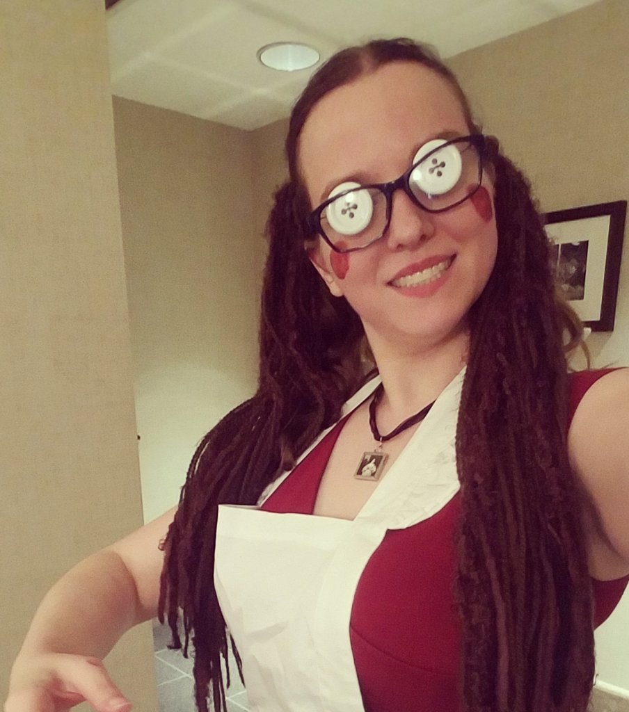 Morgan, in a red dress, covered by a white apron, dark glasses, with white button eyes. Her brown hair is in pigtails, with yarn hair-falls