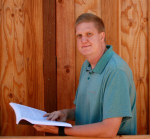 A pale, clean-faced man with short light-brown hair, wearing a blue-ish polo shirt and holding an open book in front of a wood panel background looks at the camera.