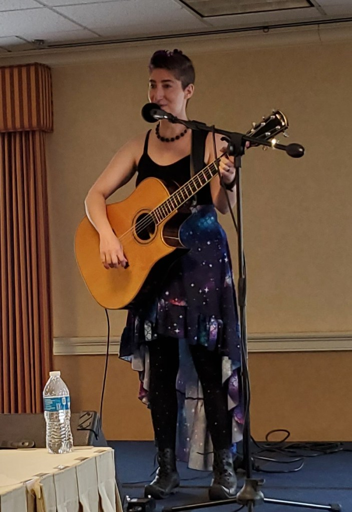 Rhiannon's Lark. Short haired, femme individual with guitar. Wearing a tank dress with a galaxy print with a high/low hemline over leggings, and black boots with upcurled toes.