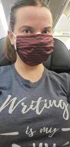 Morgan on the plane in a mask and a shirt that reads "Writing is my " (Jam)