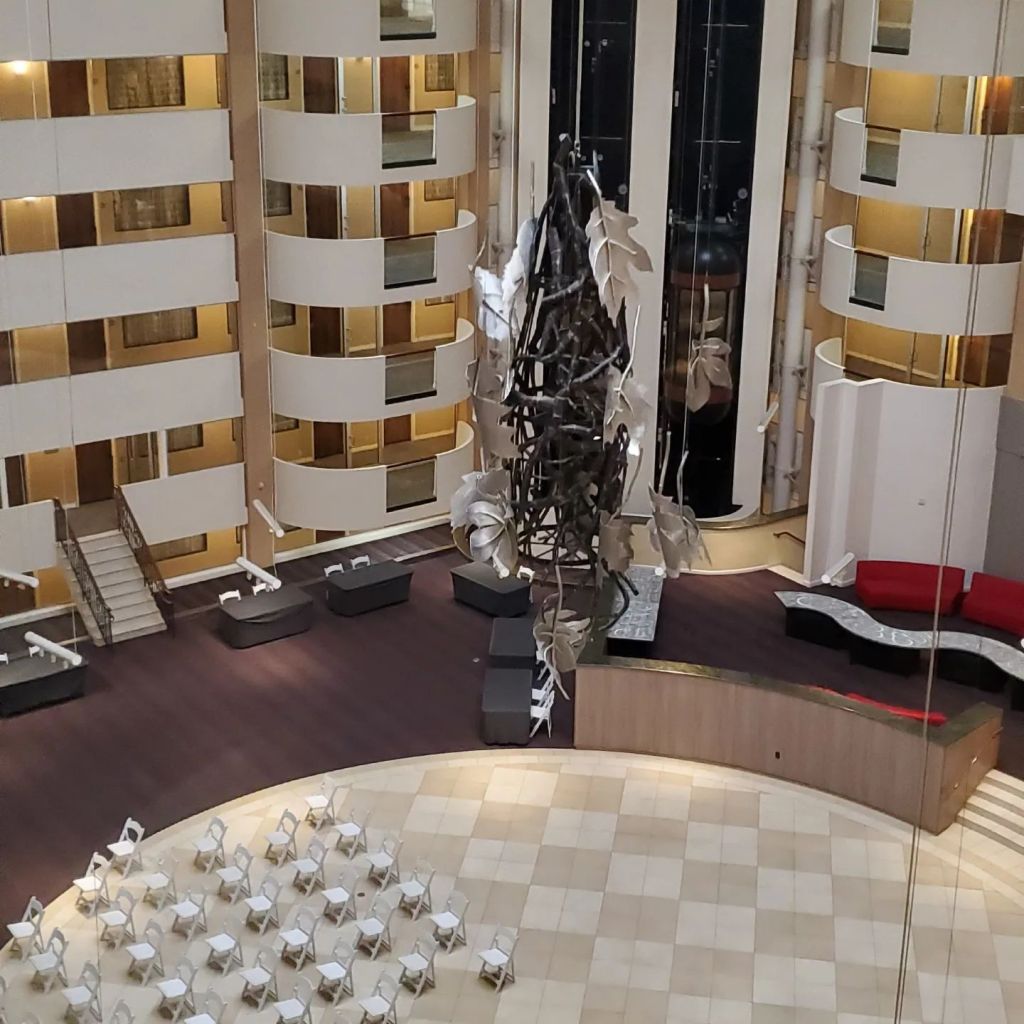Picture of a silver leaf art piece suspended in a hotel atrium, with tables at the perimeter and folding chairs lined up in rows in the center.
