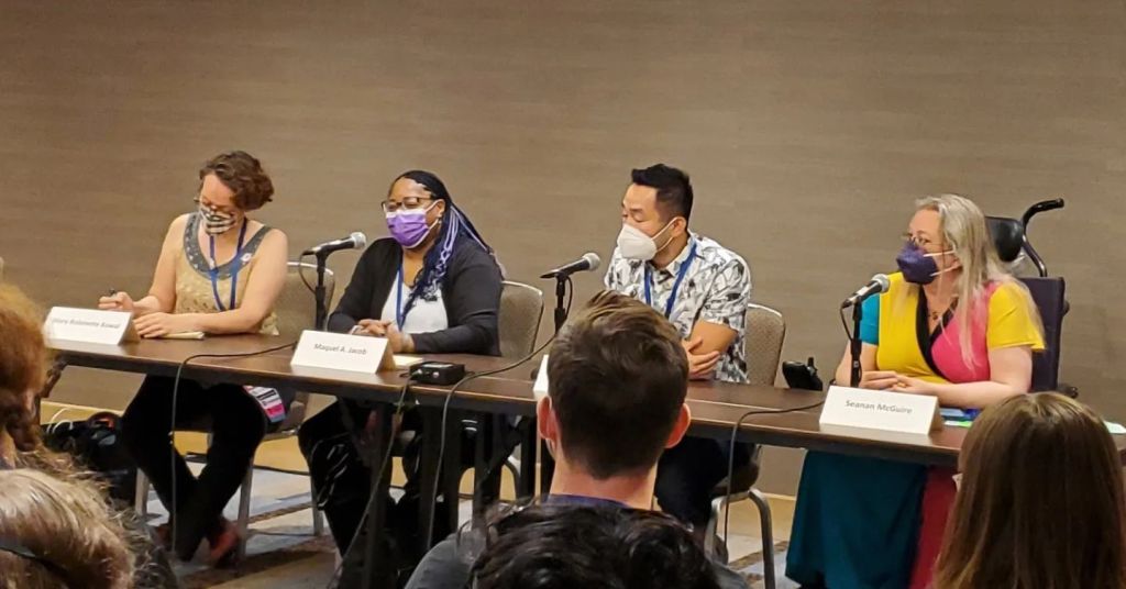 Table with microphones and 4 masked people behind it. Left to right: Mary Robinette Kowal, Maquel A Jacob, Westley Chu, and Seanan McGuire.
The backs of a few audience members heads.