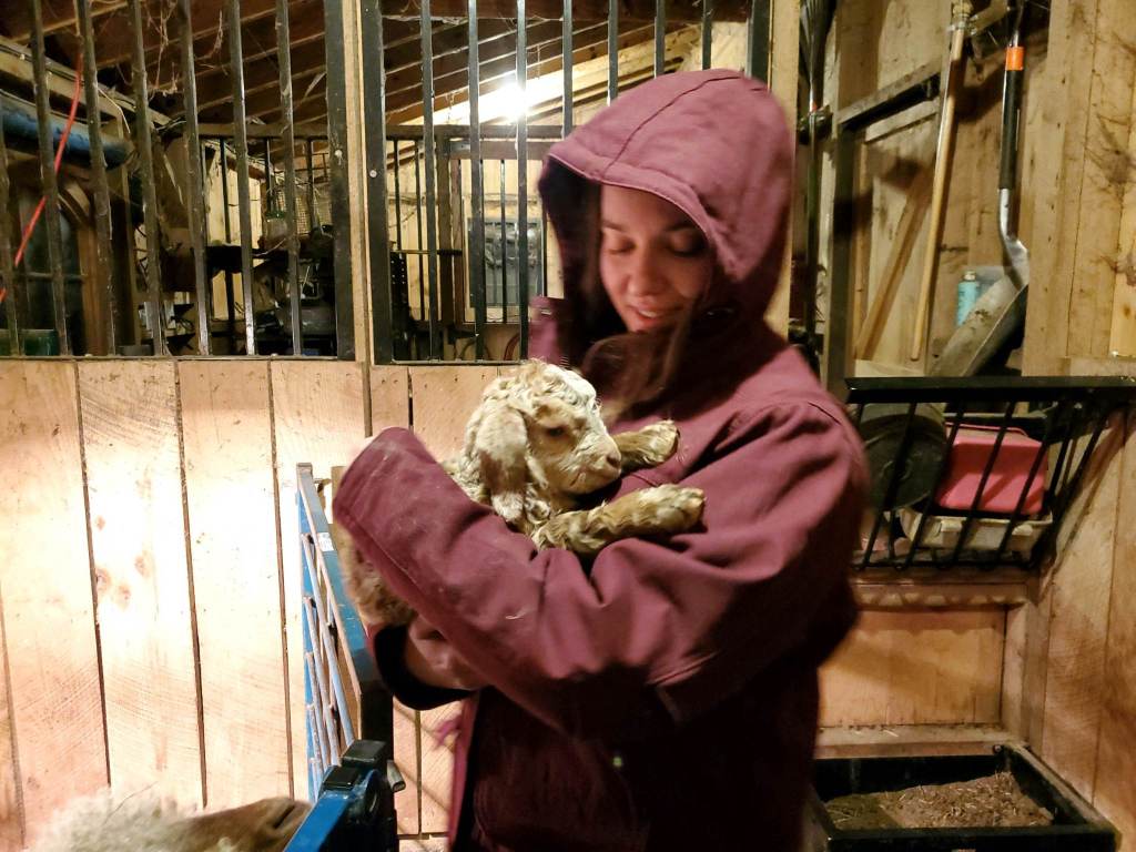 Morgan, in a red coat, holds a tiny cream colored baby goat with blond bangs. A barn surrounds them.