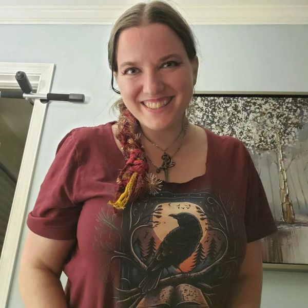 Morgan, a brunette white woman smiles at the camera, her hair hair braided with red yarn falls, wearing a dagger necklace and a burgundy shirt, with a crow sitting on a branch, next to a book.